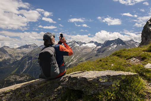 Gray-haired male traveler with backpack sitting on rock and taking pictures of mountain landscape, Austria