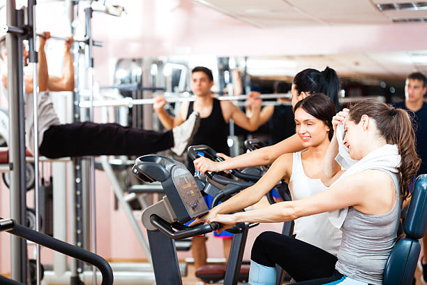 Group of young people exercising in gym. Men lifting weights, woman exercising on gym bicycle. Personal trainers assisting.