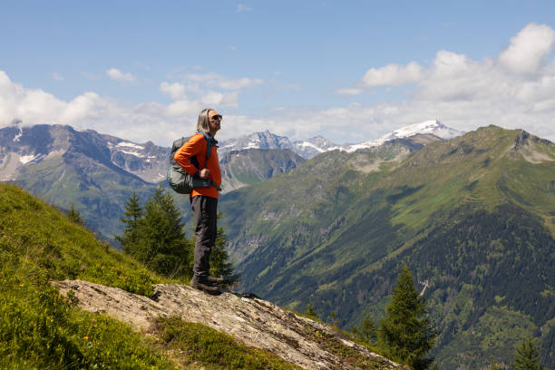 istock Gray-haired long-haired man standing on rock ledge and looking at stunning mountain view with snow-capped peaks, Austria 1754472295