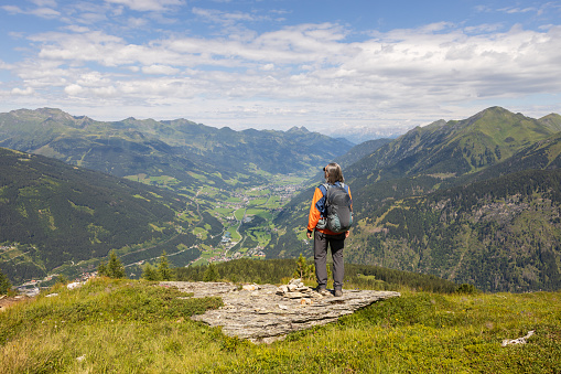 Gray-haired long-haired male hiker with backpack standing at the edge of a cliff and looking at a mountain valley, Austria. High quality photo
