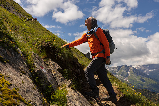 Sporty man with backpack standing on steep trail against bright cloudy sky and looking up, Austria. High quality photo