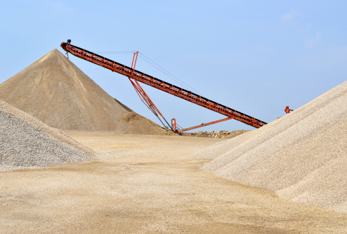 A quarry with three large mounds of recycled crushed rock with a conveyor in the background and blue sky