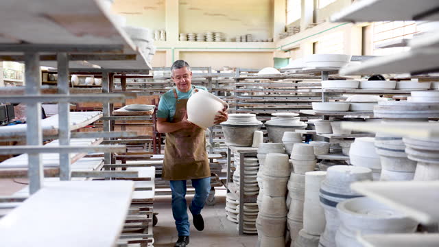 Cheerful man at a pottery factory carrying a big vase walking in between the shelfs