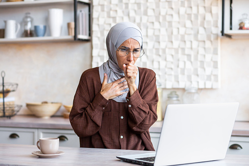Young Muslim woman in hijab is working, studying at home in the kitchen at the laptop and not feeling well. He coughs, covers his mouth with his hand, feels chest pain.