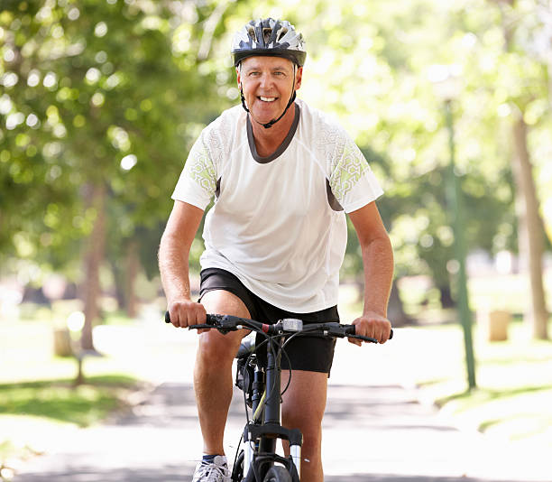 Senior man riding bike in park on sunny day Mature Man Cycling Through Park Smiling At Camera During Summer old man on bike stock pictures, royalty-free photos & images