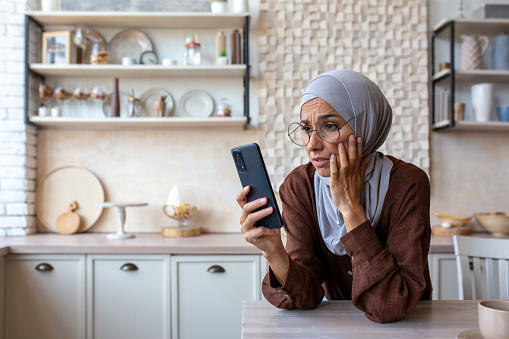 Upset and worried young muslim woman in hijab standing in the kitchen, leaning her hand on the table and looking at the phone.