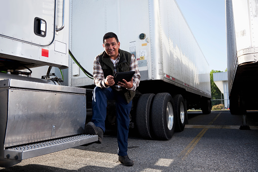 Hispanic truck driver (40s) standing next to semi-truck with digital tablet.
