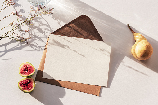 Envelope with blank card, glass of water, pear, figs and flowers in sunlight on white table. Top view, flat lay, mockup.