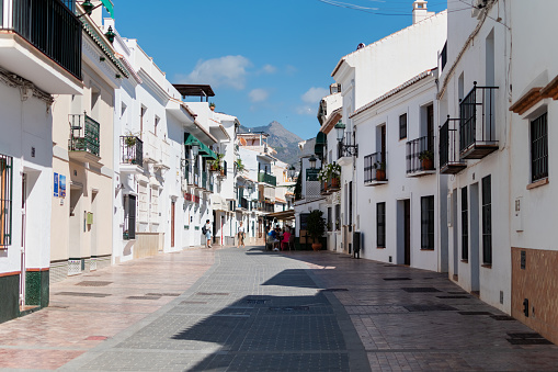 Paved pedestrian street with white houses of old town against backdrop of mountains and blue sky. Spain, Nerja, September 26, 2023