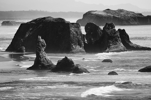 Windswept Pacific seascape of sea stacks, islands, waves and salt air, in monochrome.  Horizontally composed with a wide band of the upper center frame filled with sea stack rock and small rock islands.  A sliver at the top of frame is salt-air-filled sky.  A narrow band at the bottom of frame is water and wave action.