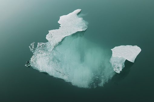 A single melting iceberg floats atop a perfectly placid water surface that appears green from sunlight refracting off pulverized glacial rock.  The iceberg is close-up, centered in the horizontal frame, and unusually shaped.  Image colors are white and green.