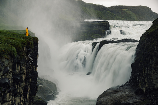 A wild nature in Iceland. A man in yellow raincoat is standing on a cliff and enjoying the view. An explorer is looking at the waterfall.