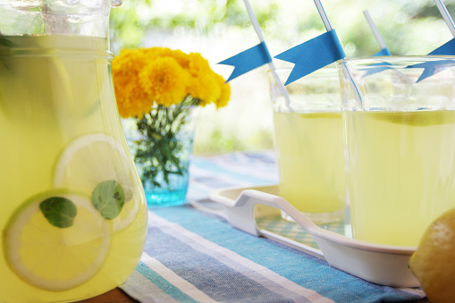 A DSLR photo of lemonade drinks in a table setting for a party at the garden. A glass jar with lemonade with mint and sliced lemons stands on the foreground at the left. Several glasses served with lemonade and decorated with blue pastel stands on the tray. The tray features a white and paste blue check pattern on top. A bouquet with yellow flowers in a blue glass vase stands behind. The table is dressed with a blue and white striped tablecloth. The garden is shown defocused at the background.