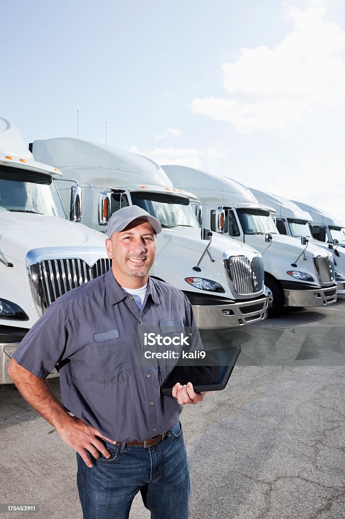 Truck driver in front of big rigs with digital tablet A truck driver standing in front of a row of parked semi-trucks, looking at the camera, holding a digital tablet.  He is wearing a gray shirt, hat and blue jeans.  He is smiling and confident, standing with his hand on his hip.  He is a mid adult man, in his 30s. Fleet of Vehicles Stock Photo