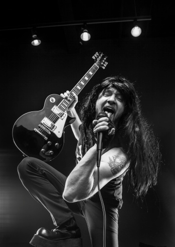 This is a close up image of a rock star with a guitar in one hand and a microphone in the other.  He is starring into the camera singing and his booted foot is raised.  There are stage lights in the background.