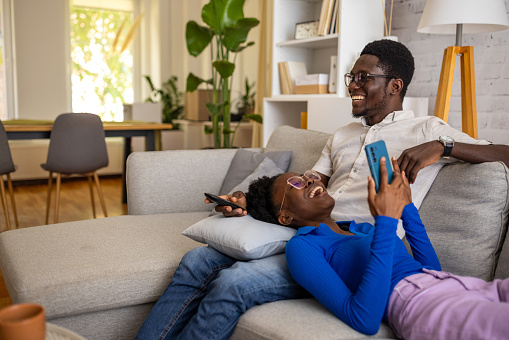 Two people, young black couple relaxing together on sofa at home. Man is watching TV, woman is using smart phone.