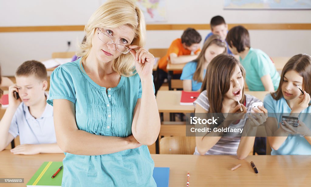 Frustrated  teacher in classroom, pupils  behind her are not listening. Disinterested pupils in classroom are not listening to the teacher. Boys are using their mobile phones and girls are putting on their make-up during lesson. Teacher Stock Photo