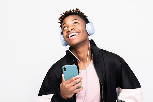 Handsome africam american guy in stylish casual clothes outfit standing over white background n studio isolated smiling holding cell phone like microphone in hands singing wearing wireless headphones.