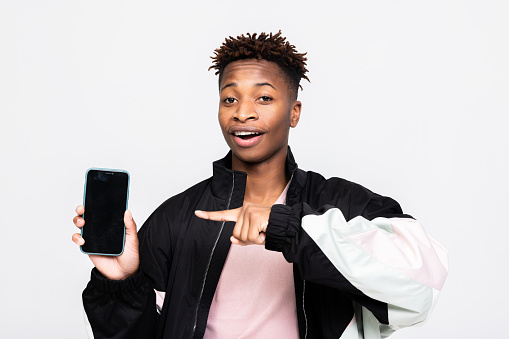 Curious inquisitive young dark skinned man guy wearig stylish new clothes looking at camera holding cell phone smartphone in hands pointing indicating at black empty screen over white background.