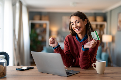 Happy young female shopper holding credit card, using laptop doing online banking transaction while sitting at desk at home.