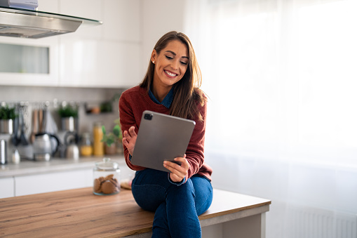 Smiling woman sitting on kitchen counter at home working on tablet pc. Professional businesswoman looking at tablet screen distantly working from home.