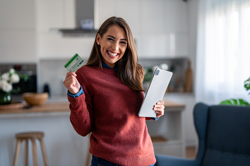Satisfied young woman looking at camera and smiling while showing credit card and holding digital tablet pc.