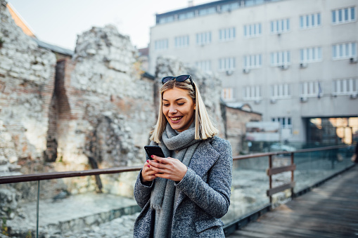Caucasian female using her phone while taking a walk in the city. Smiling young woman walking along city street texting on mobile phone.
