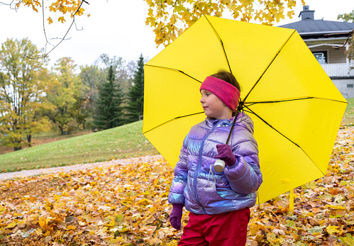 An 8-year-old girl is taking a walk in the park in the fall, holding a bright yellow umbrella in her hand and looking into the distance, thoughtful.