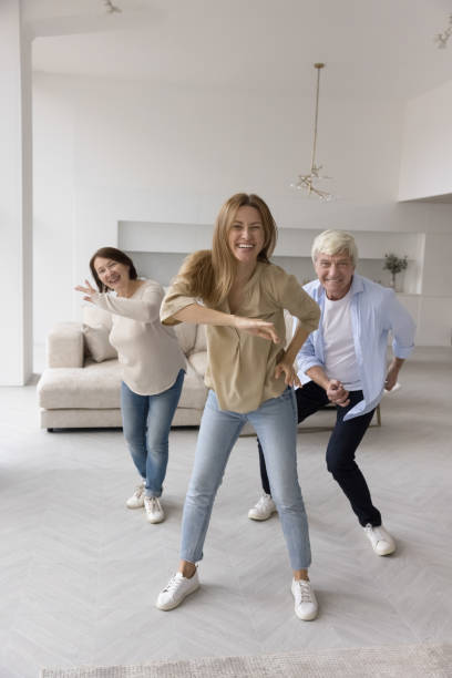Happy excited adult daughter and joyful active senior parents dancing Happy excited adult daughter and joyful active senior parents dancing, looking at camera, smiling, laughing, having fun. Trainer training elder couple at home, enjoying activity to music in apartment teacher retirement party stock pictures, royalty-free photos & images