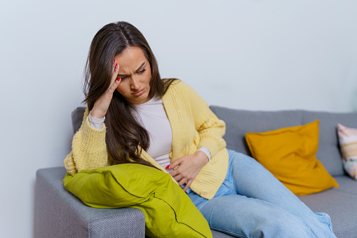 Young Caucasian woman experiencing menstrual cramps and stomach pain, while sitting on the sofa and resting her head on her hand. She has a worried lok on her face, while holding her hand on her stomach to try and reveal herself from the pain.