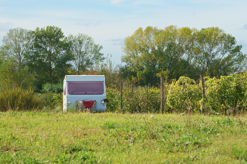 image shows a small trailer parked in the middle of a large field. The trailer is white with a green trim, and it has a single window on the side. The field is green and lush, and there are a few trees in the background. The sky is blue and cloudless. Copy space on the grass