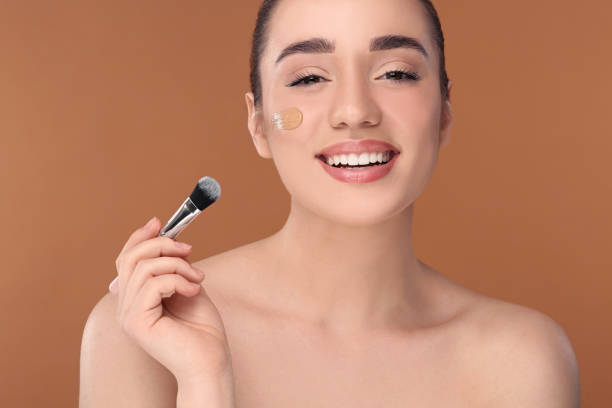 istock Woman applying foundation on face with brush against brown background 1754762830