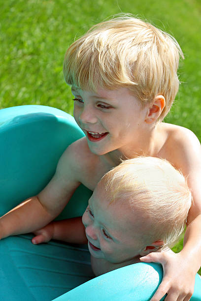 Brothers Playing on Slide Outside close up of two children: a baby and his big brother, laying at the top of a playground slide together, laughing and smiling on a summer day top of water slide photos stock pictures, royalty-free photos & images
