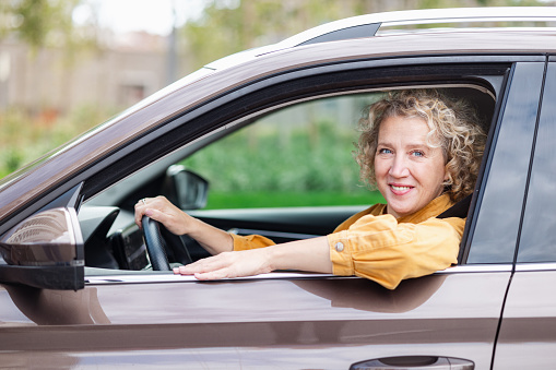A cheerful woman in her 50s is driving a car and looking at the camera through the car window