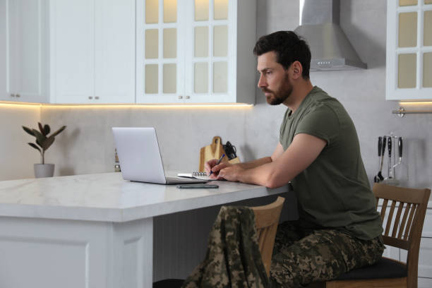 istock Soldier taking notes while working with laptop at white marble table in kitchen. Military service 1754785644
