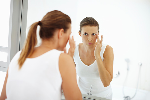 A pretty young woman applying lotion to the skin around her eyes in the mirror
