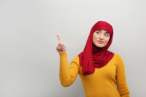 Muslim woman in hijab pointing at something on light gray background, space for text