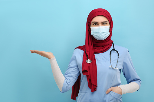 Muslim woman in hijab, medical uniform and protective mask pointing at something on light blue background. Space for text