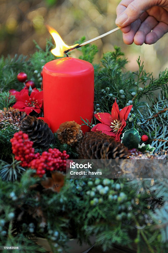 fire on Advent wreath with red candle and ornaments "Sitting a candle to burn with a matchstick. Traditional advent wreath with pine twig, red candle, pine cone and other ornaments. Winter decoration for table. Soft focus. My own hand." Advent Wreath Stock Photo