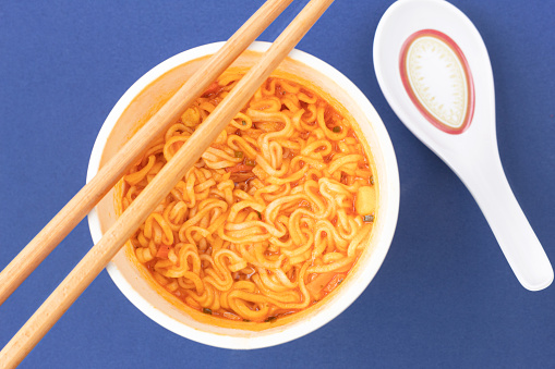 Overhead view of a bowl of instant pasta soup with a pair of chopsticks and a soup spoon on a blue background. Lunch. Fast food concept.