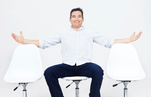 Happy adult male sitting on a white chair surrounded by empty seats with his arms stretched out