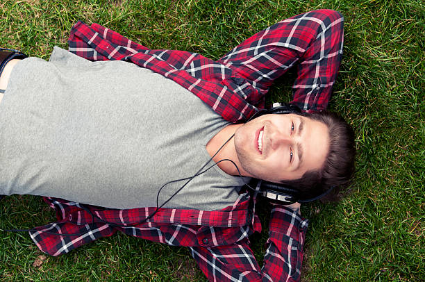 Young man is relaxing in the grass listening to music Young man wears headphones and listens to music, a podcast or an audio book. He is lying down on his back in the grass in a park. His hands are behind his head.  headphones plugged in photos stock pictures, royalty-free photos & images