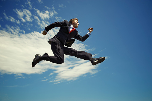Young energetic businessman jumping high outdoors leap across blue sky with clouds