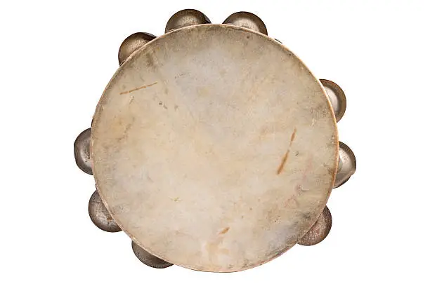 Photograph of a tambourine taken from above.  The tambourine is round and has nine jingles on its sides.  The jingles are grouped in threes, and only half of each is visible in the picture.  The tambourine is beige in color and has some signs of discoloration on its surface.  The tambourine is darker on the edges and lighter in the center.  The jingles are a dull metallic color.  On the right side of the tambourine is a brown stain that runs down in a vertical line.  The background of the image is white.