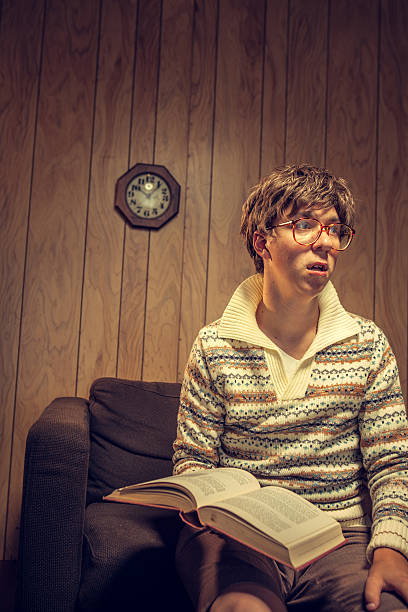 Nerd Student Studying in Vintage Room A retro styled nerd young man studies a large text book in a an arm chair, sitting in a 1970's style wood paneled living room.  He wears a gaudy sweater and shorts.  A wooden clock is on the wall behind him.  He looks like he's just read something sad or confusing.  Vertical with copy space. boring homework twelve stock pictures, royalty-free photos & images