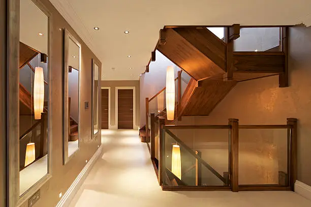 "the first floor landing in a luxurious new home with beautifully crafted woodwork stairs and stairwells. Glass panels have been used below the banisters. Three long mirrors on the wall to the left reflect the rightside view. Long lampshades are suspended from the second floor down the stairwell, creating a warm light for this scene.Please see my other Interior and Architectural images by clicking on the Lightbox link below...A>AA>A"