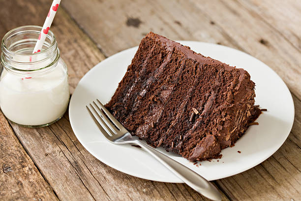 A high angle close up of a plate with a slice of chocolate fudge cake and a glass container of milk with a a straw with little red hearts on it. Shot on a grungy old wooden table.