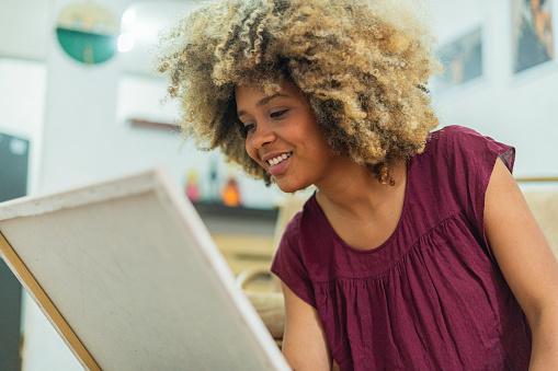 The 25-year-old Latina woman dressed comfortably with afro hair is at home painting with paints on a canvas while enjoying time alone