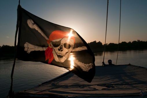 An Egyptian felucca captain stands at the back of his vessel, which is flying a pirate flag, at sunrise
