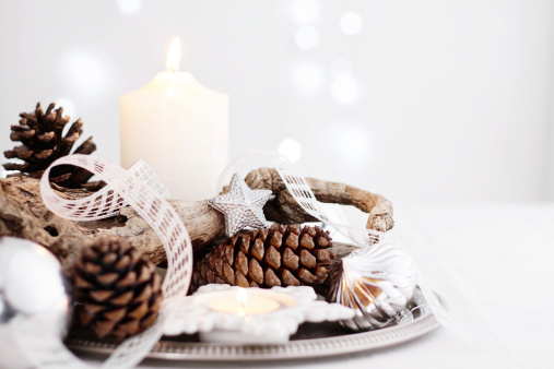 Close up of a beautiful centrepiece arranged for Christmas holidays on a table with white tablecloth. The decoration features a beautiful shaped dry branch, pine cones, white candles, silver Christmas ornaments and a delicate white bow on a silver tray. Defocused light background with Christmas lights.